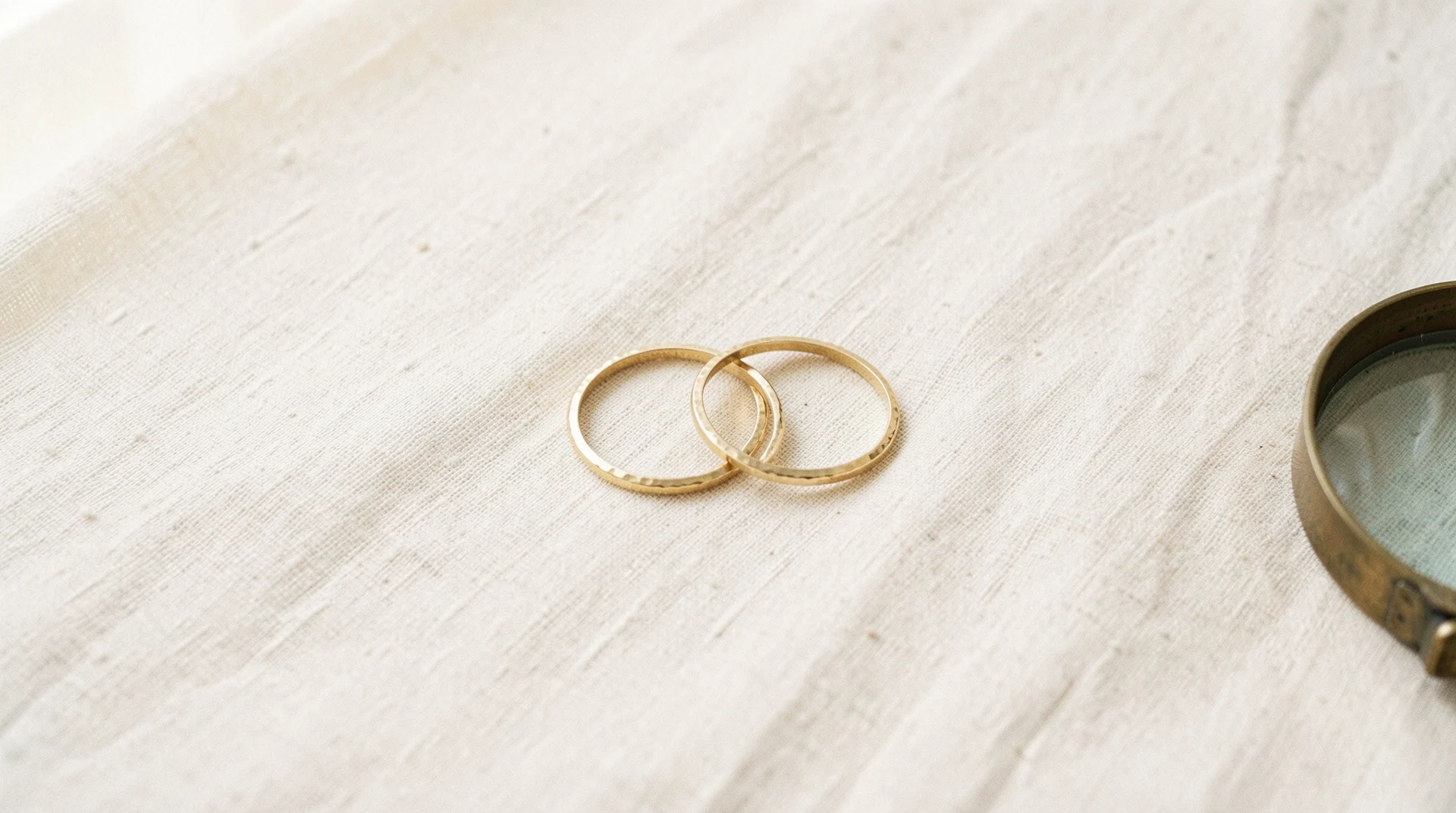 Two thin hammered gold bands, one resting on the other, on undyed linen beside a brass loupe
